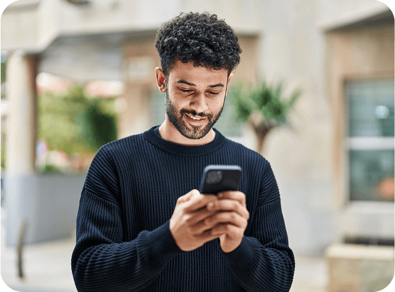 A man standing outdoors, smiling while looking at his smartphone, suggesting he is engaged in a pleasant digital interaction or reading something enjoyable.