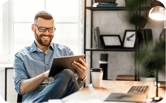 A smiling man in glasses sitting at a desk, using a tablet in a bright, modern home office, suggesting relaxed and productive remote work.