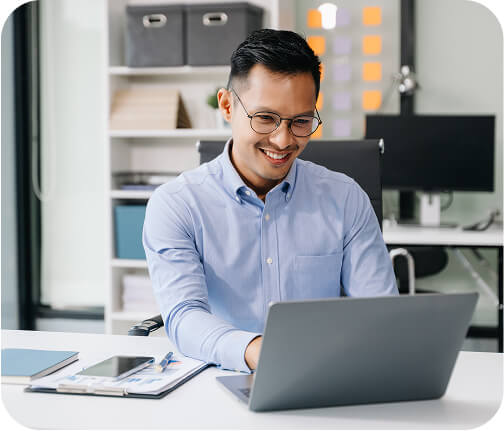 A smiling man in glasses and a light blue shirt working on a laptop at a desk in a modern office, indicating a positive and focused work atmosphere.