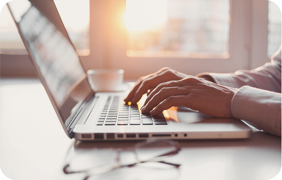 Close-up of hands typing on a laptop keyboard near a window with sunlight streaming in, suggesting focused work in a calm, bright environment.
