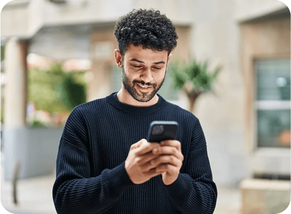 A man standing outdoors, smiling while looking at his smartphone, suggesting he is engaged in a pleasant digital interaction or reading something enjoyable.